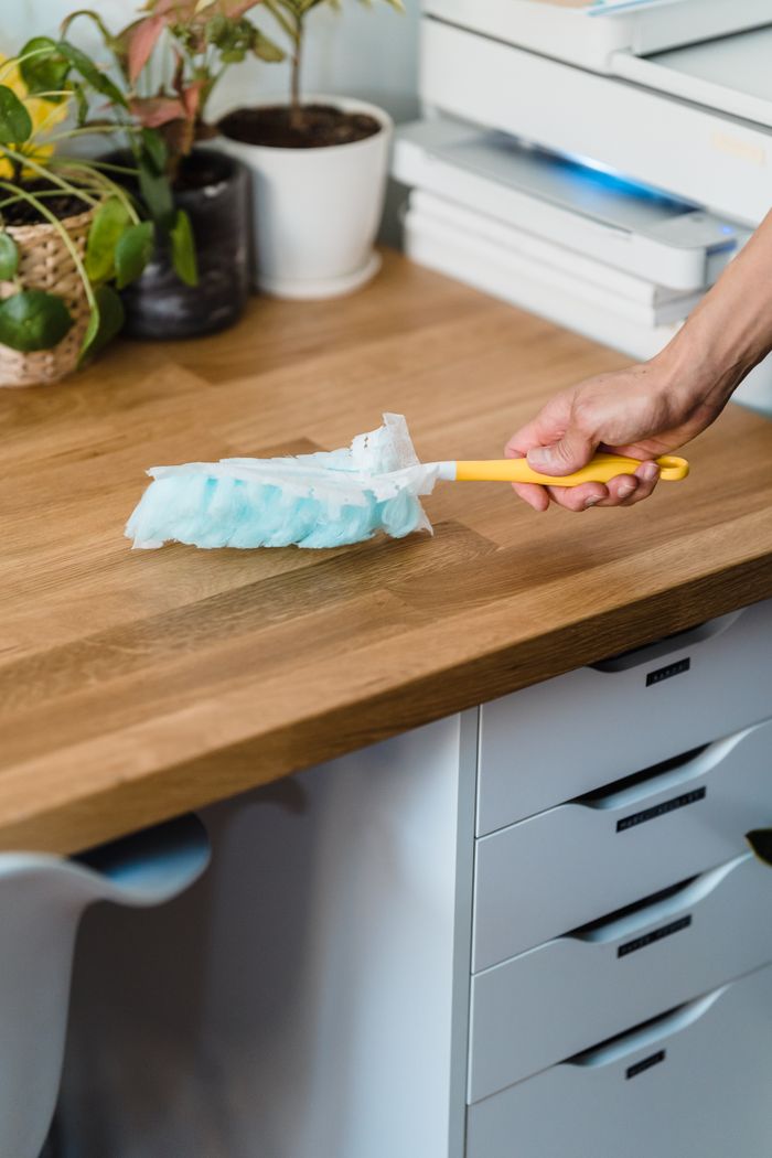 who-we-are A hand using a feather duster to clean a wooden desk, surrounded by plants and office items.